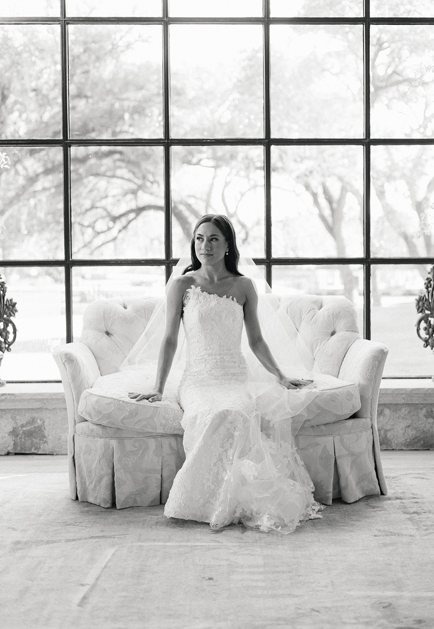 Black & white photo of bride sitting on plush couch in front of large window during bridal photoshoot at Houston Oaks Country Club